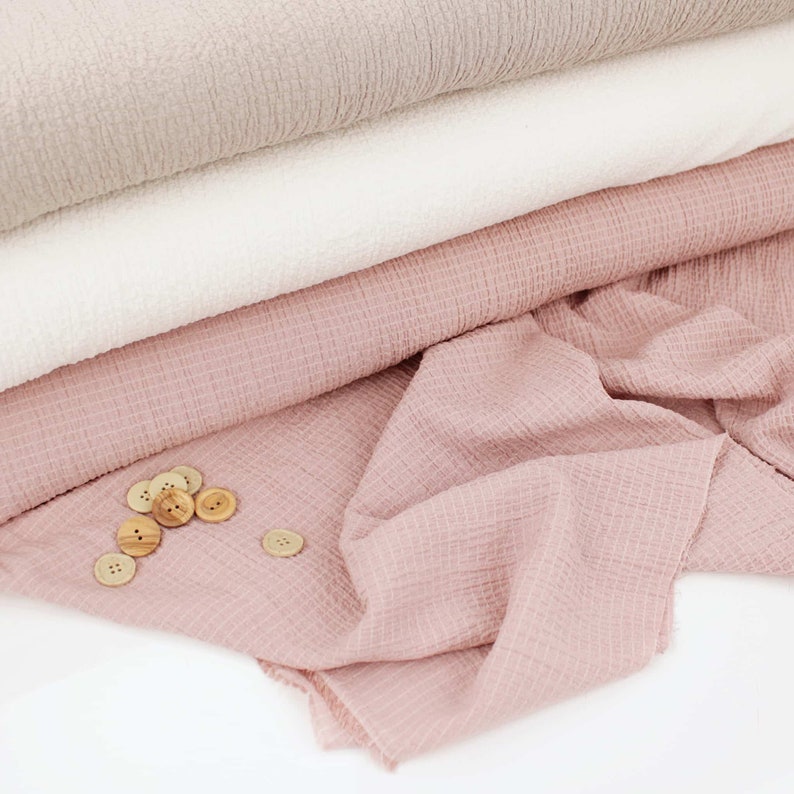 K&ouml;nnte beinhalten: Ein Stapel aus drei verschiedenen Stoffproben. Die oberste Probe ist hellbeige, die mittlere Probe ist wei&szlig; und die unterste Probe ist hellrosa. Auf dem rosa Stoff liegen mehrere Holzkn&ouml;pfe verstreut.