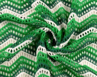 May include: Close-up of a green, white, and dark green chevron-patterned knit fabric. The fabric has a loose, open weave, creating a textured appearance. The design features alternating rows of green and white chevrons, with a darker green accent. The fabric is likely used for clothing or home decor.