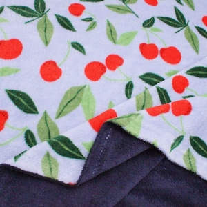 Könnte beinhalten: Weißer Stoff mit einem Muster aus roten Kirschen und grünen Blättern. Der Stoff ist gefaltet und zeigt eine dunkelgraue, plüschige Unterseite. Das Design zeigt ein sich wiederholendes Muster aus Früchten und Blättern.