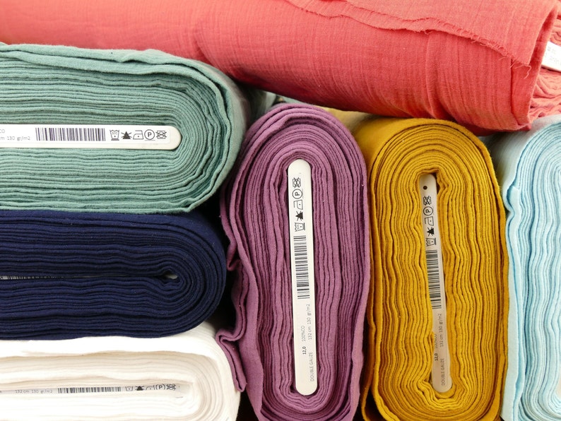 Pu&ograve; includere: Rotoli di tessuto in vari colori, tra cui verde acqua, blu navy, bianco, viola, giallo e azzurro. Ogni rotolo ha un'etichetta bianca con testo nero e un codice a barre.