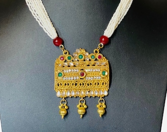 May include: A gold-toned necklace with a rectangular pendant, adorned with red, green, and clear gemstones. The pendant hangs from a multi-strand white beaded chain, with red and green beads at the top.