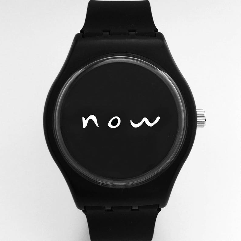 Puede incluir: Un reloj de pulsera negro con una correa de silicona negra y una esfera negra. La esfera tiene una inscripci&oacute;n blanca que dice "now".