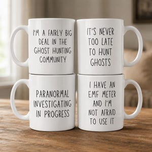 May include: Four white ceramic mugs stacked in a two-by-two grid. Each mug features black sans-serif text. The top left reads I'M A FAIRLY BIG DEAL IN THE GHOST HUNTING COMMUNITY. The top right reads IT'S NEVER TOO LATE TO HUNT GHOSTS. The bottom left reads PARANORMAL INVESTIGATING IN PROGRESS. The bottom right reads I HAVE AN EMF METER AND I'M NOT AFRAID TO USE IT.