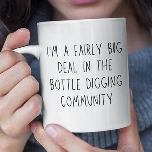 May include: White ceramic mug with black text that reads "I'm a fairly big deal in the bottle digging community."