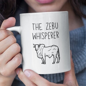 Puede incluir: Taza de cerámica blanca con un contorno negro de una vaca y el texto "The Zebu Whisperer".