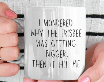 May include: White ceramic mug with black text that reads "I wondered why the frisbee was getting bigger, then it hit me".