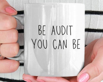 May include: White ceramic mug with the text "BE AUDIT YOU CAN BE" in black, held by a person wearing a black and white striped top. The mug has a standard handle and a smooth surface.