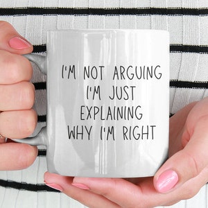 Op de afbeelding: Witte keramische mok met de tekst "I'M NOT ARGUING I'M JUST EXPLAINING WHY I'M RIGHT" in het zwart. De mok wordt vastgehouden door een persoon die een zwart-wit gestreept shirt met lange mouwen draagt. De mok is een standaard formaat.