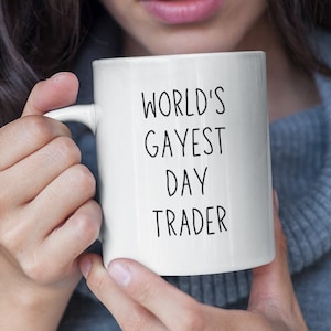 May include: A white ceramic mug with black text that reads "World's Gayest Day Trader". A person is holding the mug.