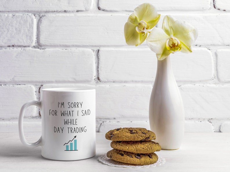 May include: White ceramic mug with a black graphic that reads "I'm sorry for what I said while day trading" with a blue upward trending line graph. The mug is on a white surface with a vase of white orchids and chocolate chip cookies.