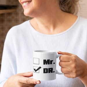 Op de afbeelding: Witte keramische mok met een zwarte checklist met de tekst "Mr." en "DR." met een vinkje naast "DR."