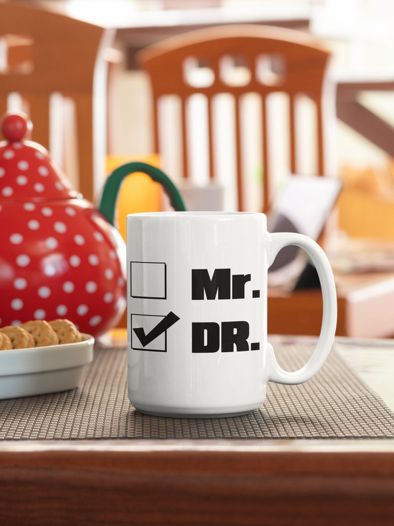 Op de afbeelding: Witte keramische mok met een zwart vinkje en de tekst "Mr." ernaast. Een tweede zwart vinkje is aangevinkt en de tekst "DR." staat ernaast.