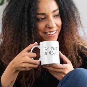 May include: White ceramic coffee mug with black text that reads "I get paid to argue". A person is holding the mug and smiling.