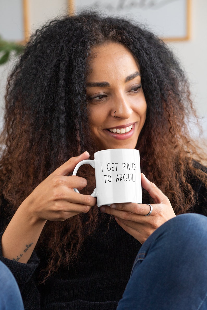 May include: White ceramic coffee mug with black text that reads "I get paid to argue". A person is holding the mug and smiling.