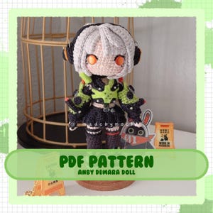 Puede incluir: Un patrón de amigurumi de crochet para un personaje llamado Anby Demara. La muñeca lleva un atuendo negro y verde con ojos naranjas. El patrón está disponible para descargar en PDF.