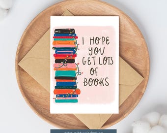 May include: A greeting card with the text "I HOPE YOU GET LOTS OF BOOKS" and a stack of colorful books with string lights. The card is on a wooden tray with a brown envelope.
