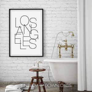 May include: A black and white print with the word "LOS ANGELES" in a stylized font, framed in black, hanging on a white brick wall. A white bathtub with gold fixtures is in the foreground.