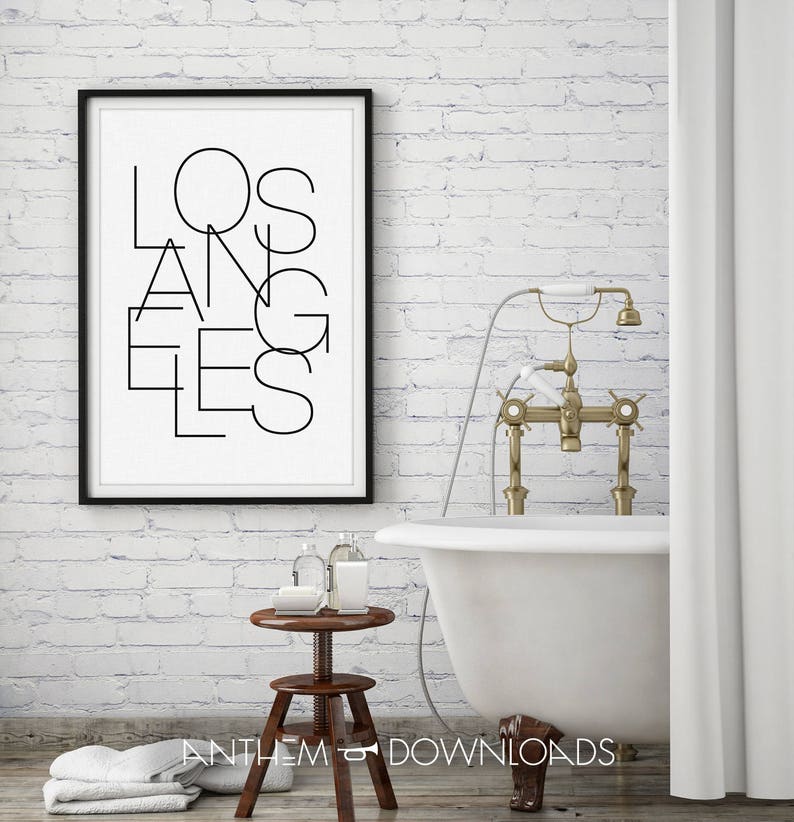 May include: A black and white print with the word "LOS ANGELES" in a stylized font, framed in black, hanging on a white brick wall. A white bathtub with gold fixtures is in the foreground.