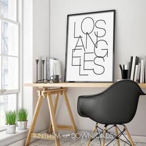 May include: A black and white print with the word "LOS ANGELES" in a modern, geometric font. The print is framed in a black frame and is hanging on a white wall above a wooden desk.