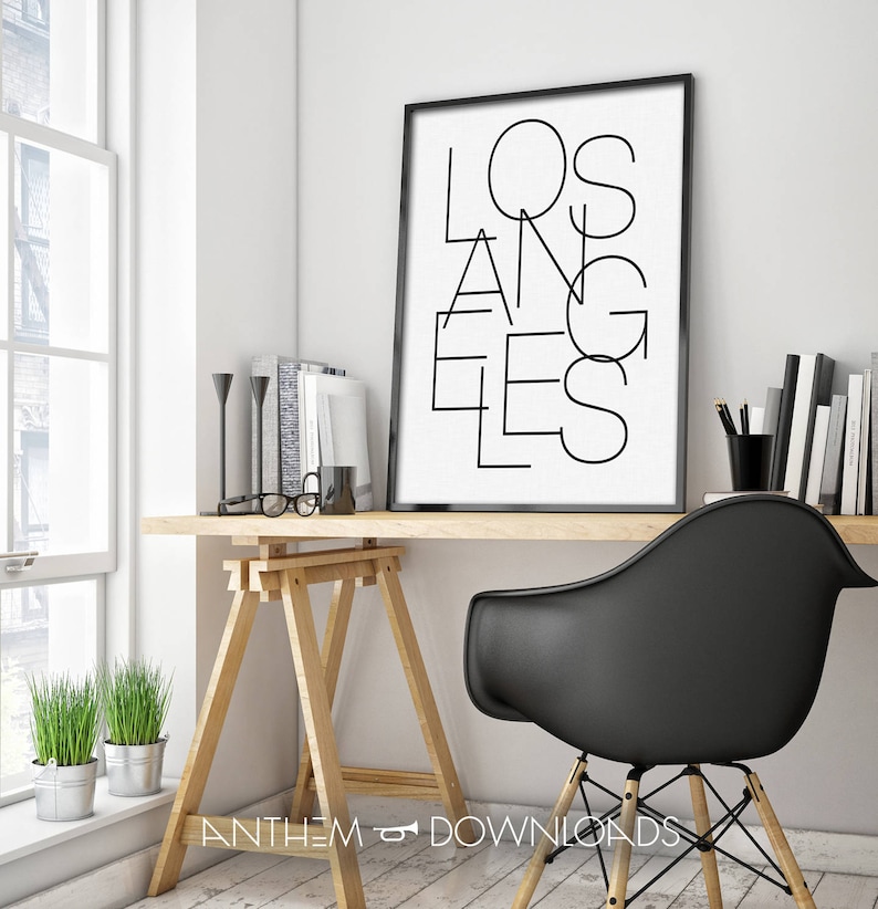 May include: A black and white print with the word "LOS ANGELES" in a modern, geometric font. The print is framed in a black frame and is hanging on a white wall above a wooden desk.