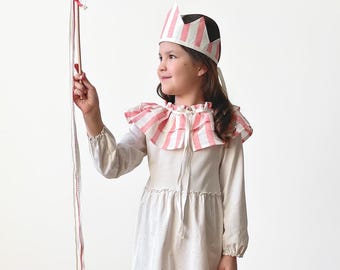 May include: A child's costume featuring a white dress with a ruffled collar and a matching crown, both accented with pink stripes. The child is holding a wand with pink and white ribbons. The outfit is completed with white knee-high socks.