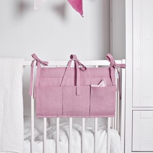 May include: A pink fabric organizer with multiple pockets hangs on a white crib. The organizer has tie closures and is filled with small items. A white waffle-weave blanket is draped over the crib rail. A white wardrobe is in the background.