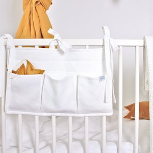 May include: White fabric crib organizer with multiple pockets, attached to the crib with ties. A gold star-shaped toy is in one of the pockets. A white crib and bedding are in the background.