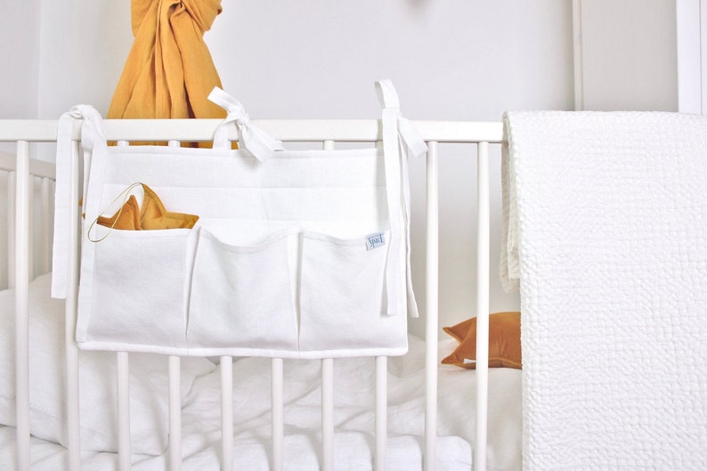 May include: White fabric crib organizer with multiple pockets, attached to the crib with ties. A gold star-shaped toy is in one of the pockets. A white crib and bedding are in the background.