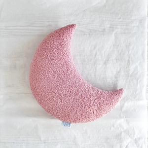 May include: A pink crescent moon-shaped pillow with a textured surface. The pillow has a small blue tag that reads "Space". The pillow is lying on a white surface.