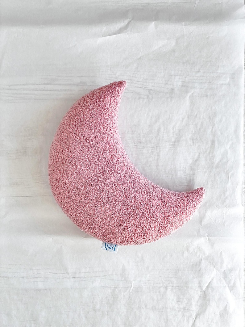 May include: A pink crescent moon-shaped pillow with a textured surface. The pillow has a small blue tag that reads "Space". The pillow is lying on a white surface.