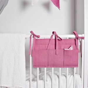 May include: A pink nursery organizer with multiple pockets hangs on a white crib. A white textured blanket is draped over the crib. Pink bows secure the organizer to the crib. A white cabinet is visible in the background.