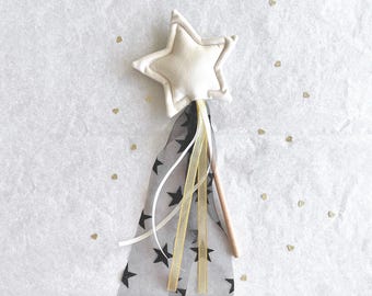 May include: A cream-colored star-shaped wand with a wooden handle. The wand features a black and gray star-patterned fabric with gold and white ribbons. Gold heart-shaped confetti is scattered around the wand.
