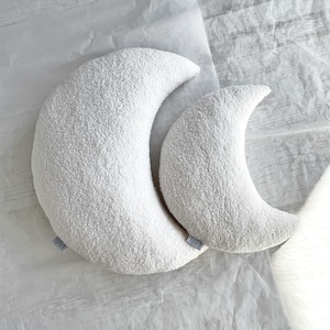 May include: Two white crescent moon-shaped pillows with a textured, plush surface. The pillows are different sizes and rest on a white, textured surface. Each pillow has a small, light blue tag.