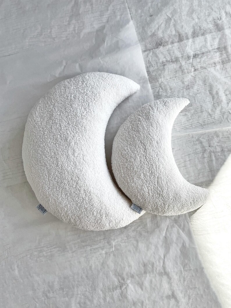 May include: Two white crescent moon-shaped pillows with a textured, plush surface. The pillows are different sizes and rest on a white, textured surface. Each pillow has a small, light blue tag.