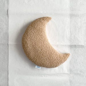 May include: A crescent-shaped, tan-colored pillow with a textured surface. The pillow has a small tag that reads "Spact". The pillow is lying on a white surface.