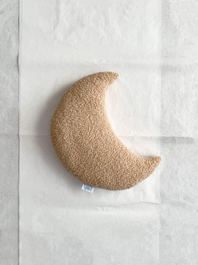 May include: A crescent-shaped, tan-colored pillow with a textured surface. The pillow has a small tag that reads "Spact". The pillow is lying on a white surface.