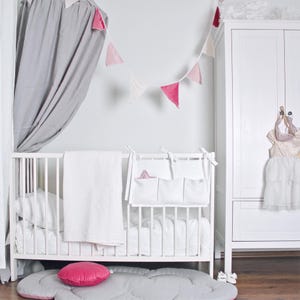 May include: A white crib with bedding, a white blanket, and a pink pillow. A white organizer with pockets hangs on the crib. A pink and white pennant banner hangs above the crib. A white wardrobe and a cloud-shaped rug complete the nursery.