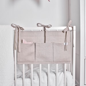 May include: A beige linen crib organizer with three pockets and tie closures. The organizer hangs on the crib and has a soft, neutral color. A white blanket is visible on the left side of the image.