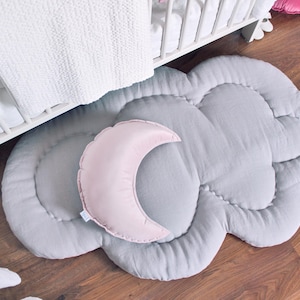 以下が含まれることがあります： ピンクの三日月がアクセントになった、グレーの雲型のフロアマット。柔らかいキルティング生地で作られており、木製の床に置かれています。白いベビーベッドと毛布が背景に見えます。