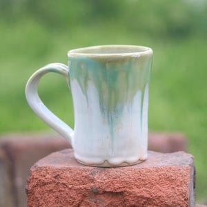 May include: A ceramic mug with a cylindrical body and a curved handle. The mug features a white base glaze with a reactive green and blue drip pattern cascading from the rim down the sides. The surface has a glossy finish with visible texture.