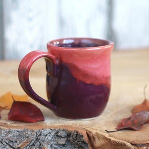 May include: A ceramic mug featuring a two-tone glaze with a deep purple base that transitions into a vibrant coral-pink upper section. The mug has a rounded body, a curved handle, and a smooth, glossy finish throughout the exterior surface.
