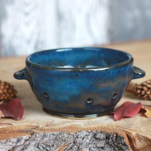 May include: A round ceramic bowl with two small side handles and a series of small circular drainage holes along the lower perimeter. The bowl features a deep blue glaze with mottled brown and dark undertones, creating a textured appearance.