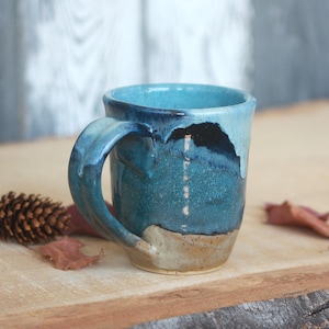 May include: A ceramic mug featuring a reactive glaze with shades of deep blue, turquoise, and a light tan base. The mug has a sturdy handle and a smooth, rounded rim. The glaze drips down the sides, creating a layered effect with dark blue accents.