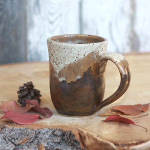 May include: A ceramic mug featuring a two-tone glaze with a speckled cream-coloured upper section and a smooth, dark brown lower section. The mug has a curved handle and a cylindrical body, showcasing a distinct transition between the two colours.