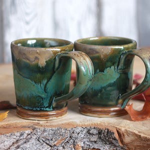 May include: Two handmade ceramic mugs featuring a glossy glaze in shades of deep teal, forest green, and earthy brown. The mugs have a cylindrical shape with a sturdy handle and a distinct drip pattern near the rim, showcasing a smooth finish.