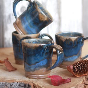 Peut inclure: Un ensemble de trois tasses en céramique avec un émail réactif aux tons bleu profond et brun terreux. Les tasses ont une forme cylindrique avec une anse robuste, présentant une finition lisse où l'émail bleu coule sur la base brune.
