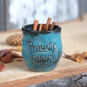 May include: A small, turquoise ceramic cup with the words "Proudly CANADA STRONG!" inscribed on the front. The cup is filled with cinnamon sticks and sits on a wooden surface, with autumn leaves scattered around.