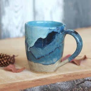 May include: A ceramic mug featuring a layered glaze design with a light beige base, transitioning into deep navy blue and bright turquoise at the top. The mug has a curved handle and a smooth, glossy finish, showcasing a distinct drip-like pattern.