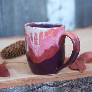 May include: A handcrafted ceramic mug with a distinctive colour gradient. The mug transitions from a deep purple base to shades of red and pink, with white drips near the rim. The handle is a matching red-purple. The mug is set on a wooden surface.