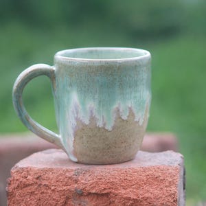 May include: A ceramic mug featuring a smooth, glossy glaze that transitions from a soft, seafoam green at the top to a speckled, earthy tan at the base. The mug has a curved handle on the left side and a cylindrical body with a slightly tapered bottom.